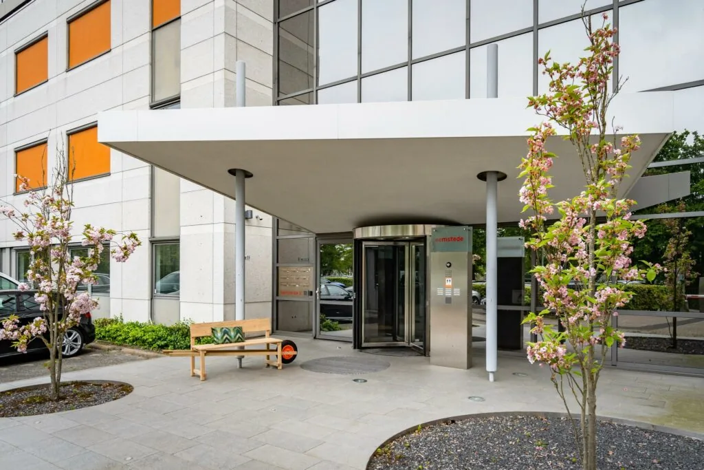 Ingang van het kantoorgebouw aan de Baarnsche Dijk met draaideur, bloeiende bomen en een houten bankje.